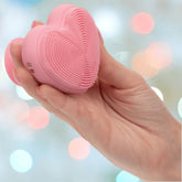 A hand holds the CalExotics Posh Kiss Me Handheld Massager with textured pleasure nubbies and a suction base against a blurred background of pastel-colored lights.